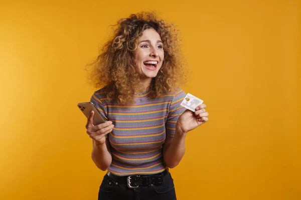 快乐的年轻女子拿着手机和信用卡庆祝在黄墙背景下的成功 — 图库照片