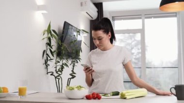 Güzel bir kadın evdeki mutfakta salata pişirirken cep telefonunu kullanıyor.