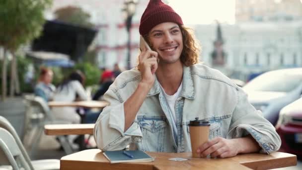 一个穿着牛仔裤夹克的快乐男人坐在外面街上的咖啡厅阳台上 一边用手机聊天 — 图库视频影像