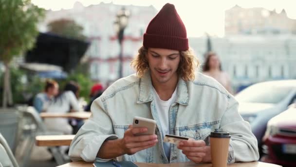 路上のカフェテラスに座ってお金を送金ジーンズのジャケットを着て幸せな男 — ストック動画