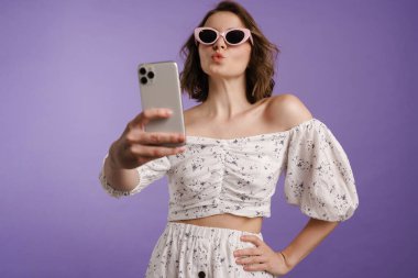 Mor arka planda cep telefonu tutan, selfie çeken, güneş gözlüğü takan mutlu beyaz kadın.