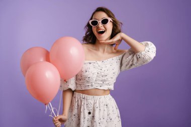 Mor duvarda izole edilmiş balonlarla poz veren heyecanlı genç bir kadın.