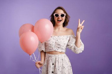 Mor duvarda izole edilmiş balonlarla poz veren heyecanlı genç bir kadın.