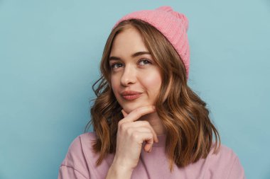 Genç beyaz kadın mavi arka planda izole bir şekilde düşünüyor ve çenesine dokunuyor.