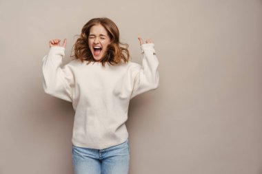 Gri arka planda izole edilmiş kameraya bağıran ve el kol hareketi yapan heyecanlı genç bir kadın.