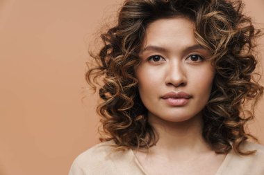 Genç kıvırcık İspanyol kadın poz veriyor ve bej arka planda izole edilmiş kameraya bakıyor.