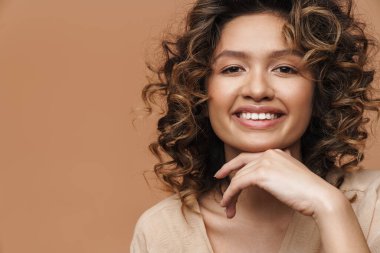 Genç kıvırcık İspanyol kadın gülümsüyor ve bej arka planda izole edilmiş kameraya bakıyor.