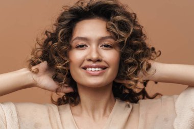 Genç, kıvırcık, İspanyol bir kadın gülümseyip saçlarına dokunuyor.