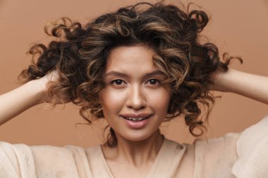 Genç, kıvırcık, İspanyol bir kadın gülümseyip saçlarına dokunuyor.