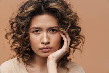 Genç kıvırcık İspanyol kadın poz veriyor ve bej arka planda izole edilmiş kameraya bakıyor.