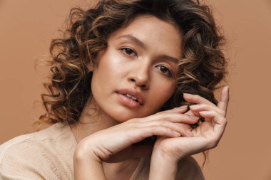 Genç kıvırcık İspanyol kadın poz veriyor ve bej arka planda izole edilmiş kameraya bakıyor.