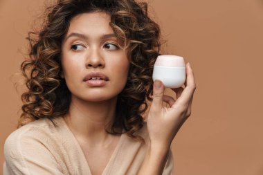 Genç kıvırcık İspanyol kadın bej arka planda izole edilmiş yüz kremi ile poz veriyor.