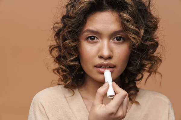 Young hispanic curly woman applying lipstick and looking at camera isolated over beige background