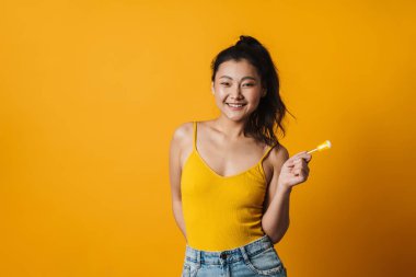 Lolipopu olan mutlu genç Asyalı kadın turuncu arka planda yazlık giysiler giyiyor.