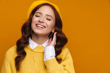 Sarı duvarın arkasında duran uzun kırmızı saçlı ve bereli mutlu genç kadın, gözleri kapalı.