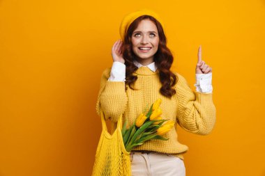 Uzun kırmızı kazaklı ve bereli gülümseyen genç bir kadın elinde lale buketi, üzerinde meyveler olan sarı duvar arkasını gösteren bir çanta taşıyor.