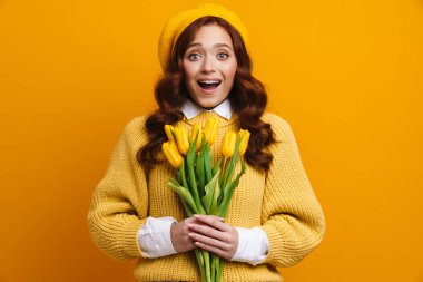 Sırıtkan, uzun kırmızı saçlı, kazaklı ve bereli genç bir kadın. Sarı arka planda lale buketi tutuyor.