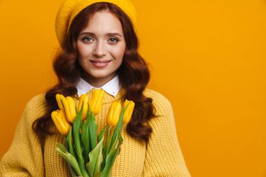 Sırıtkan, uzun kırmızı saçlı, kazaklı ve bereli genç bir kadın. Sarı arka planda lale buketi tutuyor.