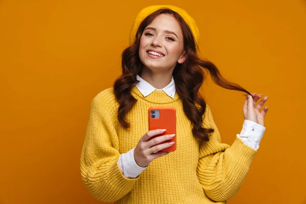穿着毛衣 头戴红色长发 头戴贝雷帽 站在黄墙后面 拿着手机 快乐而又梦幻般的年轻女子 — 图库照片
