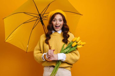 Gülümseyen kızıl saçlı kadın, sarı duvarın arkasında şemsiye altında duran laleleri tutuyor.