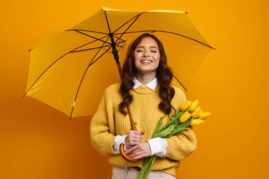 Gülümseyen, kırmızı saçlı, bereli, süveterli, elinde lale buketi ve şemsiyeyle sarı arka planda duran beyaz bir kadın.