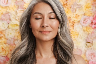 Gülümseyen olgun üstsüz kadın çiçeklerin arkasında poz veriyor, gözleri kapalı.