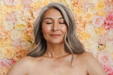 Gülümseyen olgun üstsüz kadın çiçek duvarı arkasında duruyor.