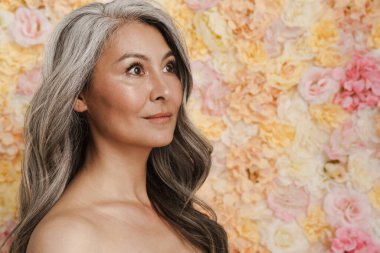 Gülümseyen olgun üstsüz kadın çiçeklerin arkasında dikiliyor, yan bakıyor.