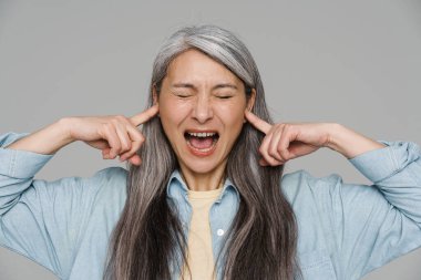 Gri duvarın üzerinden kulaklarını tıkarken çığlık atan sinirli, beyaz saçlı kadın.