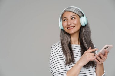 Gülümseyen beyaz saçlı kadın gri duvardan izole edilmiş cep telefonu ve kulaklıklarla müzik dinliyor.