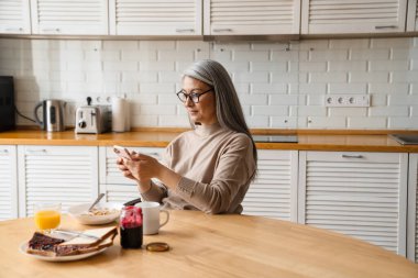 Evde kahvaltı yaparken cep telefonu kullanan olgun gri kadın.