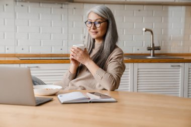 Evde dizüstü bilgisayarla çalışırken kahve içen olgun bir kadın.