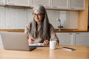 Evde dizüstü bilgisayarla çalışırken kulaklık kullanan olgun bir kadın.