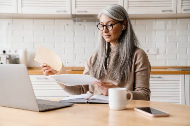 Evde dizüstü bilgisayar ve kağıtlarla çalışırken kulaklık kullanan olgun bir kadın.