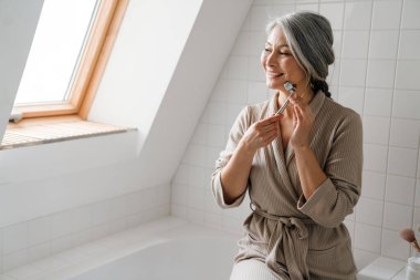 Olgun gri kadın evde tuvalette yeşim taşıyla yüzüne masaj yaparken gülümsüyor.