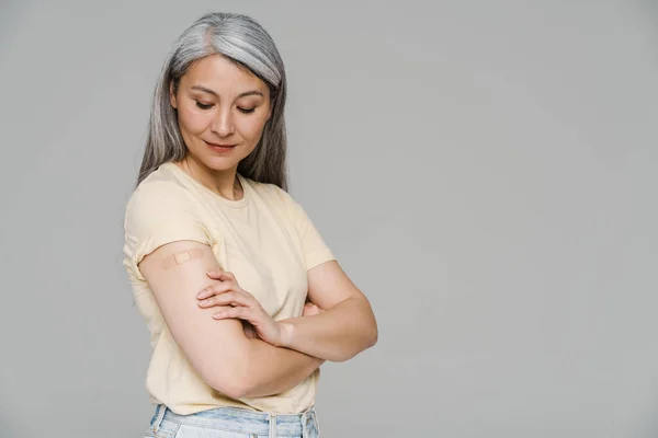一个成熟的白发女人 在灰色的墙壁上凝视着她手臂上的绷带 — 图库照片