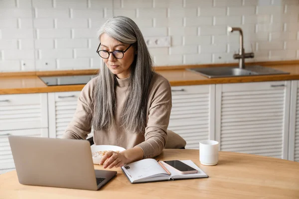 Evde mutfaktayken dizüstü bilgisayarla çalışan olgun gri kadın.