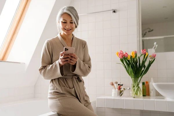 Érett Szürke Mosolyog Használata Közben Mobiltelefon Fürdőszobában Otthon — Stock Fotó