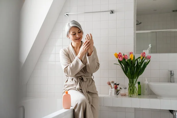Mujer Sonriente Madura Aplicando Crema Mientras Está Sentada Baño Casa — Foto de Stock