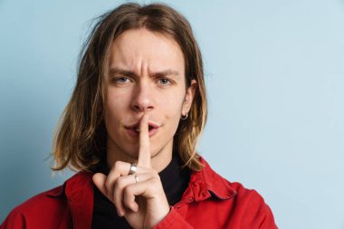 Genç beyaz adam mavi duvarın üzerinde izole edilmiş kameraya sessiz jest yaparken kaşlarını çatıyor.