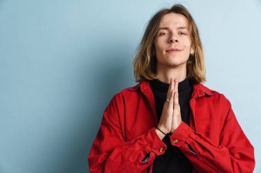 Avuç içlerini bir arada tutarken gülümseyen genç beyaz adam mavi duvarın üzerinde izole edilmiş.