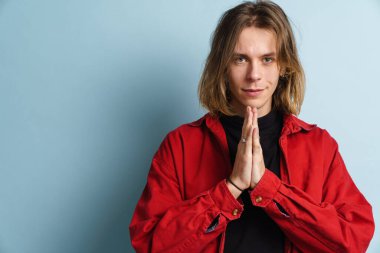 Mavi duvarın üzerinde avuç içlerini bir arada tutan genç beyaz adam kameraya bakıyor.