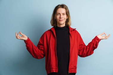 Genç beyaz adam gülümsüyor ve mavi duvarın üzerinde meditasyon yapıyor.