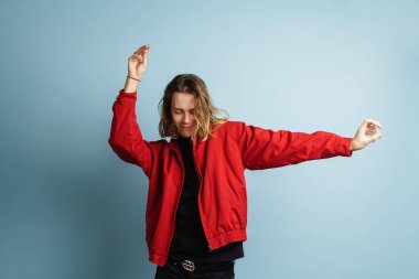 Kırmızı ceket giyen genç beyaz adam mavi duvarın üzerinde dans ediyor ve gülümsüyor.