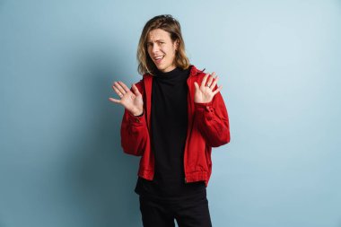 Kafası karışmış genç bir adam kameraya bakıyor ve avuçlarını mavi duvarın üzerinde soyutlanmış olarak gösteriyor.