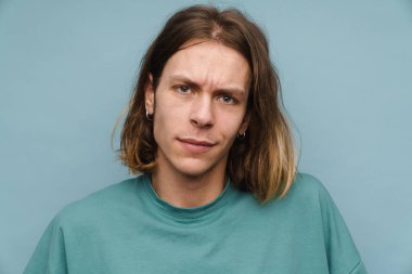 Mavi arka planda duran, kaşlarını çatmış, tişörtlü genç bir adamın portresi.