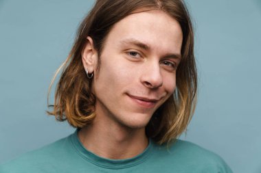 Tişörtlü gülümseyen genç sarışın bir adamın portresi mavi arka planda kameraya bakıyor.