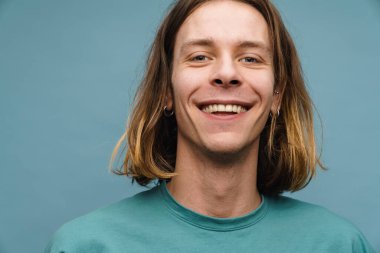 Tişörtlü gülümseyen genç sarışın bir adamın portresi mavi arka planda kameraya bakıyor.