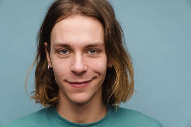 Tişört giyen genç beyaz adam gülümsüyor ve mavi duvarın üzerinden izole edilmiş kameraya bakıyor.