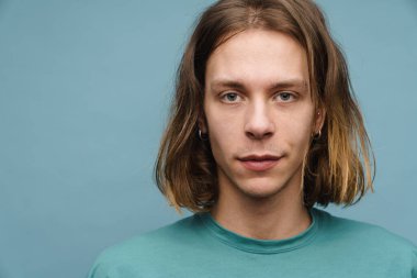 Mavi arka planda dikilip kameraya bakan tişörtlü genç sarışın bir adamın portresi.